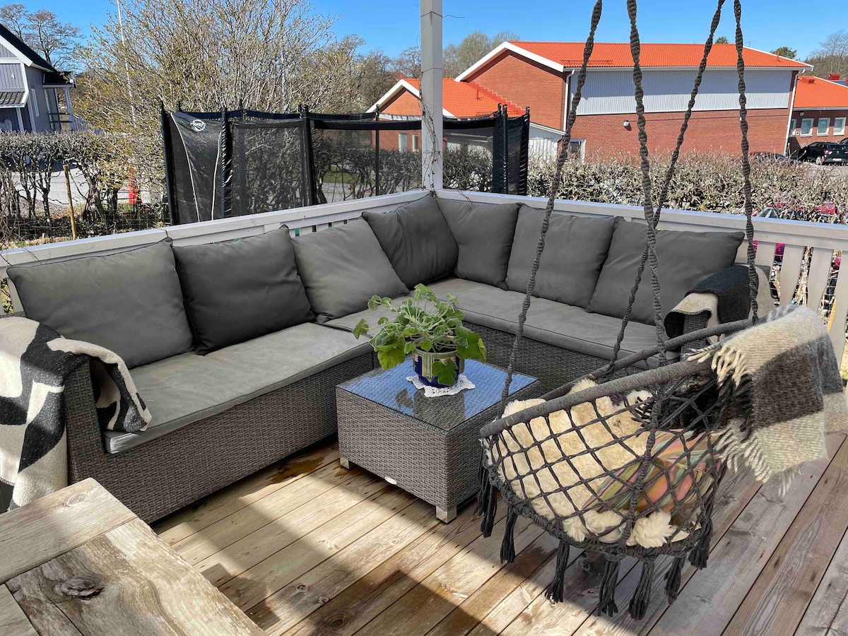 A spacious outdoor seating area features a large sectional sofa with cushions and a small table at the center. A hanging chair adds a cozy touch, surrounded by a wooden deck and a view of greenery in the background.