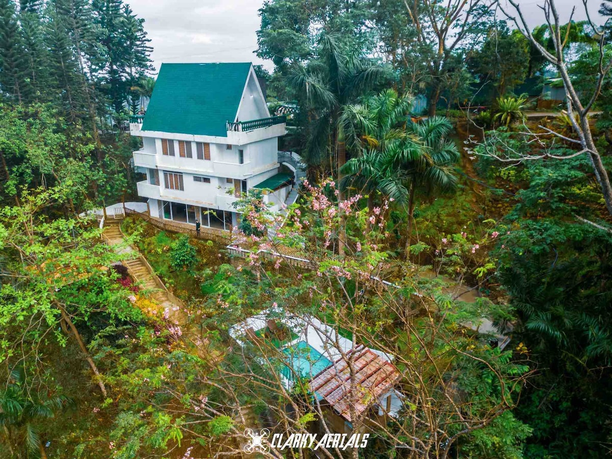 A three-story house with a green roof is surrounded by lush greenery and trees. A pathway leads to an outdoor area featuring a pool, while a cozy atmosphere is created by the surrounding nature. The overall landscape showcases a serene retreat.