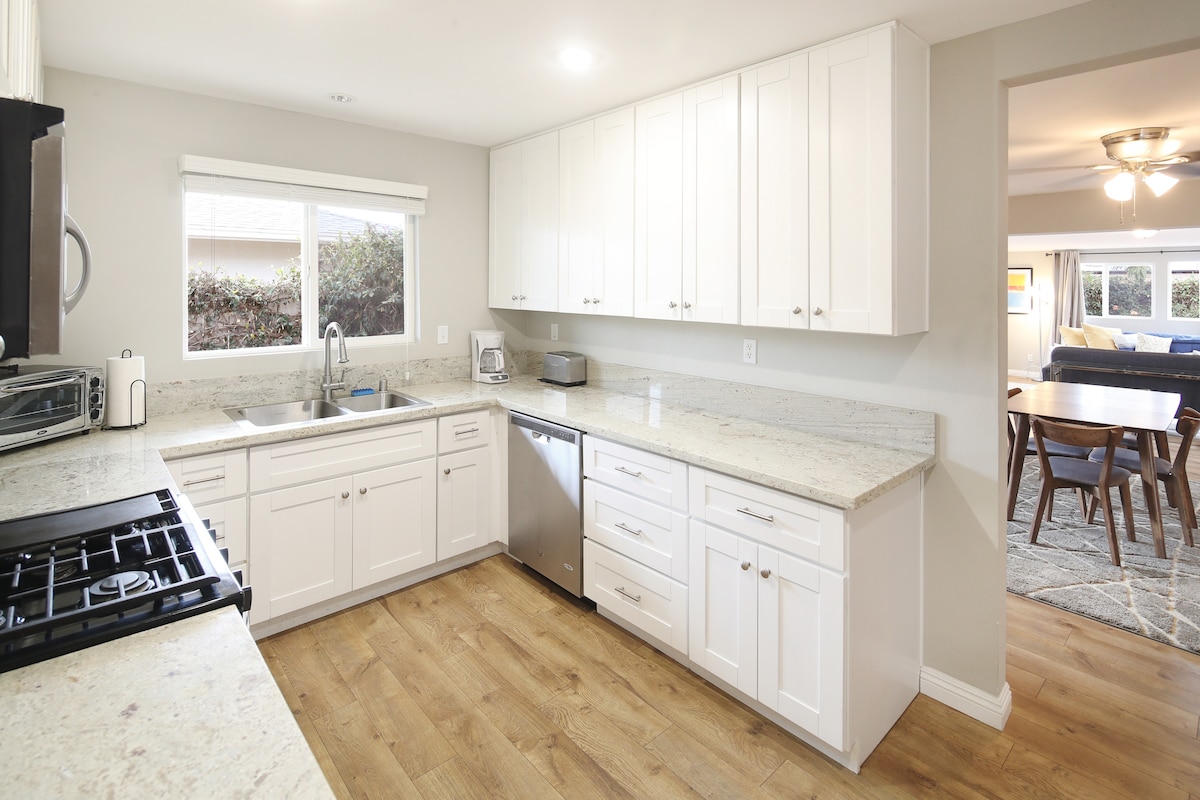 A modern kitchen features white cabinetry and granite countertops, complemented by stainless steel appliances. Natural light enters through a large window, illuminating the space. Wooden flooring extends through this area, providing warmth and continuity to the design, which opens into the dining area in the background.