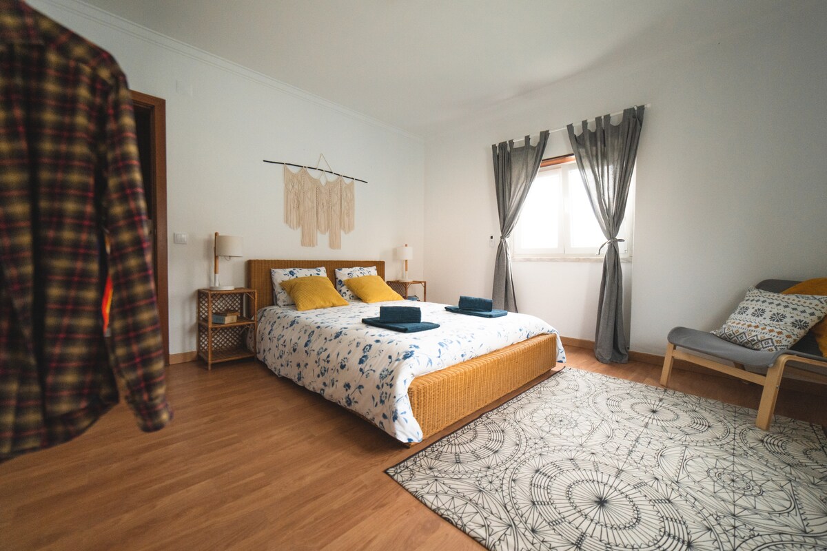 A light-filled room features a spacious bed with a floral duvet and neatly arranged towels. Soft curtains frame the window, and a patterned rug adds warmth. A chair in the corner provides additional seating, complementing the cozy, inviting environment.