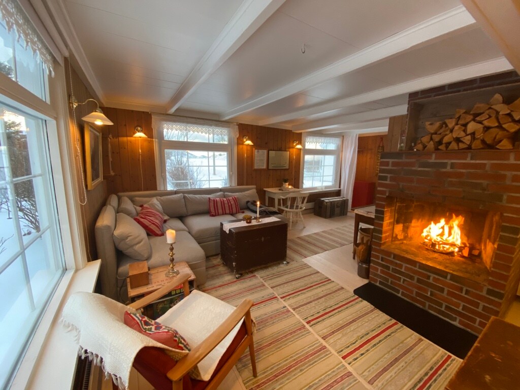 The inviting living room features a comfortable grey sofa arranged around a rustic brick fireplace, where a warm fire flickers. Large windows frame the snow-covered outdoor landscape, and a dining table sits nearby, complemented by soft lighting.