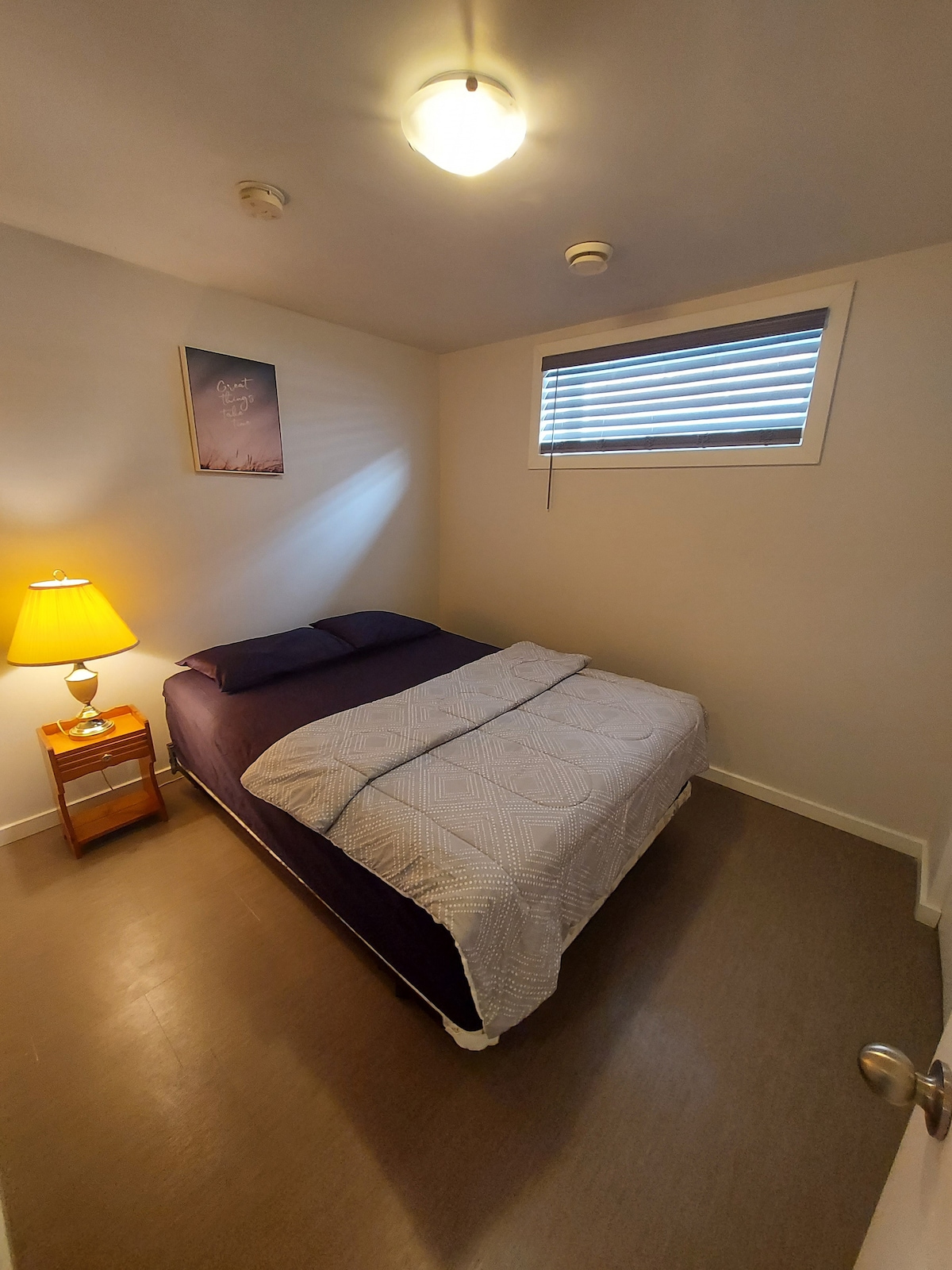 A cozy sleeping area features a spacious bed covered with a light-colored quilt and accented by purple bedding. A bedside table with a lamp provides soft lighting. A window with blinds allows natural light while maintaining privacy.