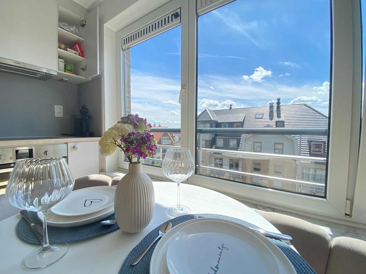 A dining area with a round table set for two is shown, featuring elegant glassware and neatly arranged plates. A vase with fresh flowers sits at the center, while large windows allow ample natural light and provide a view of the surroundings.