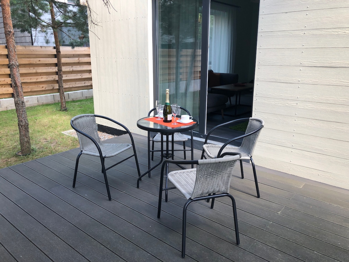 A small outdoor seating area is presented on a wooden deck. Four chairs surround a round table that holds a champagne bottle and two glasses. A glass door leads to the interior space, with greenery visible in the surroundings.