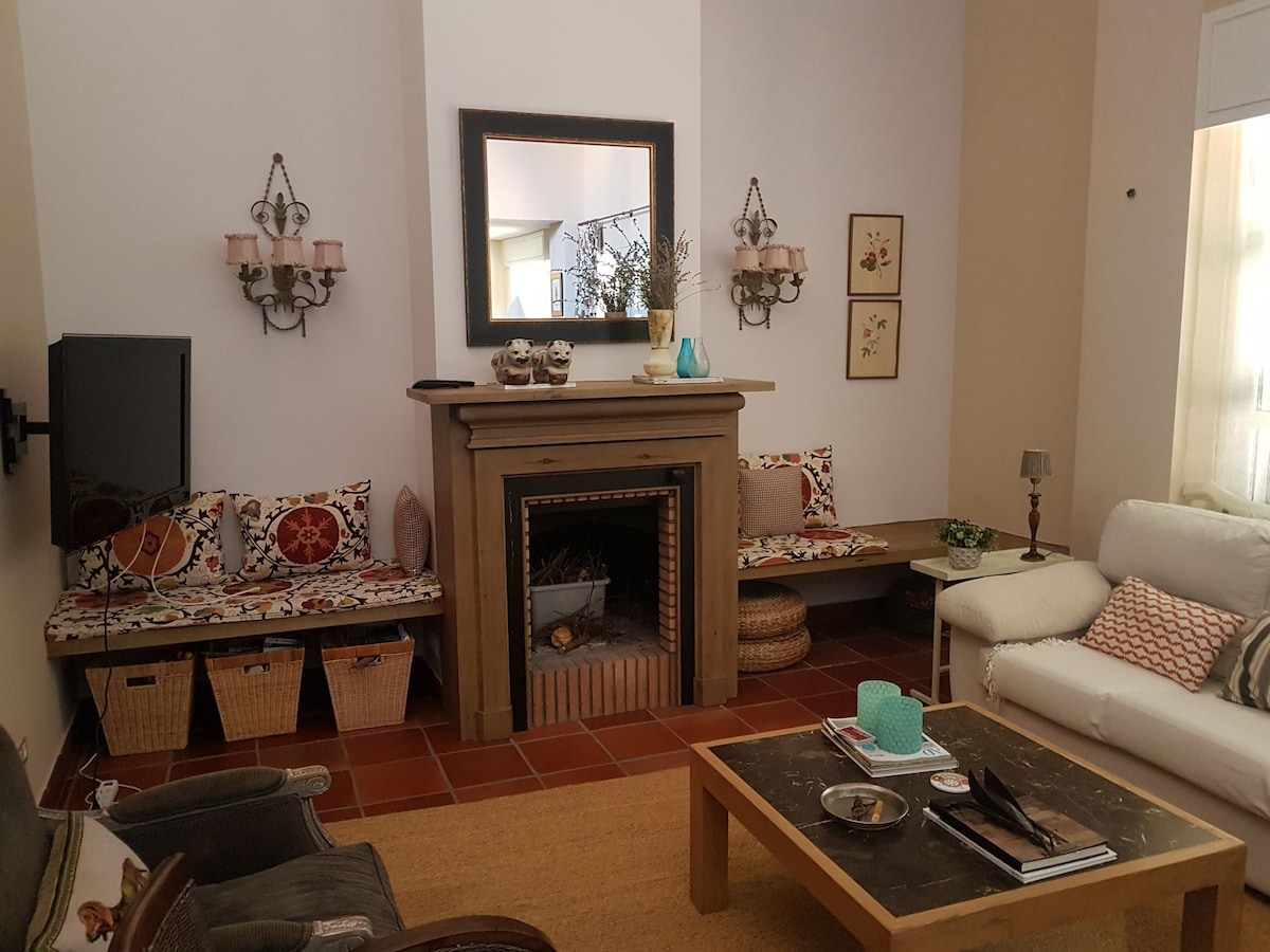The living area features a central fireplace adorned with decorative elements. Cozy seating is provided by two benches, each with patterned cushions. A flat-screen TV is mounted on the wall, and a coffee table occupies the center of the room, accompanied by a light-colored sofa.