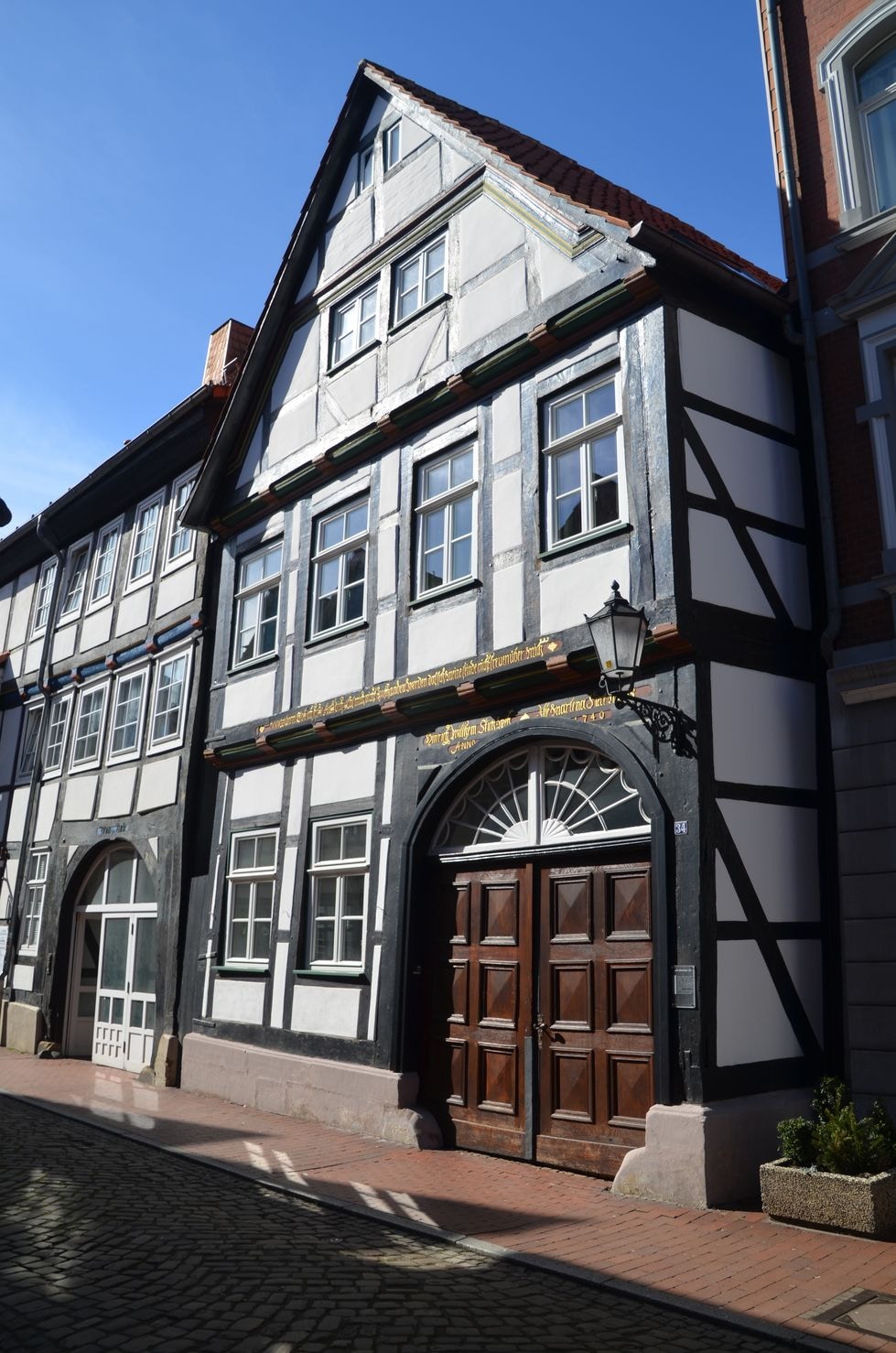The historic half-timbered building features a symmetrical facade with decorative woodwork and a large wooden door. Bright sunlight illuminates the exterior, highlighting the contrast between the black beams and white stucco. Nearby, a stone-paved street leads to a welcoming atmosphere.