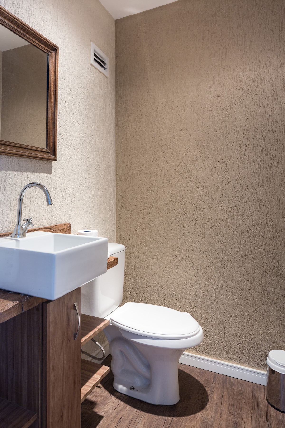 The bathroom features a wooden vanity with a square sink and a mirror above. A modern toilet is positioned adjacent to the sink. Textured beige walls complement the wood accents, and the floor is covered in dark laminate, providing a clean and organized appearance.