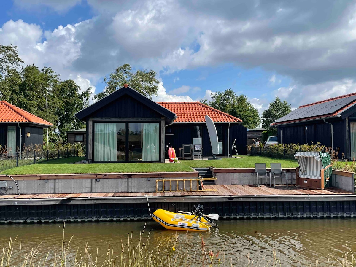 The house is presented with a dark exterior, featuring large glass doors that open to a grassy area. A small yellow boat is moored alongside a wooden dock. Sun loungers and a shaded seating area are visible on the terrace, inviting relaxation by the water.