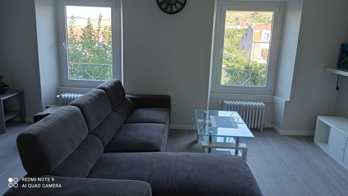 A modern living space is presented, featuring a spacious gray sectional sofa complemented by a glass coffee table. Natural light enters through two windows, illuminating the room, while a decorative wall clock is mounted above the sofa, adding a functional element to the design.
