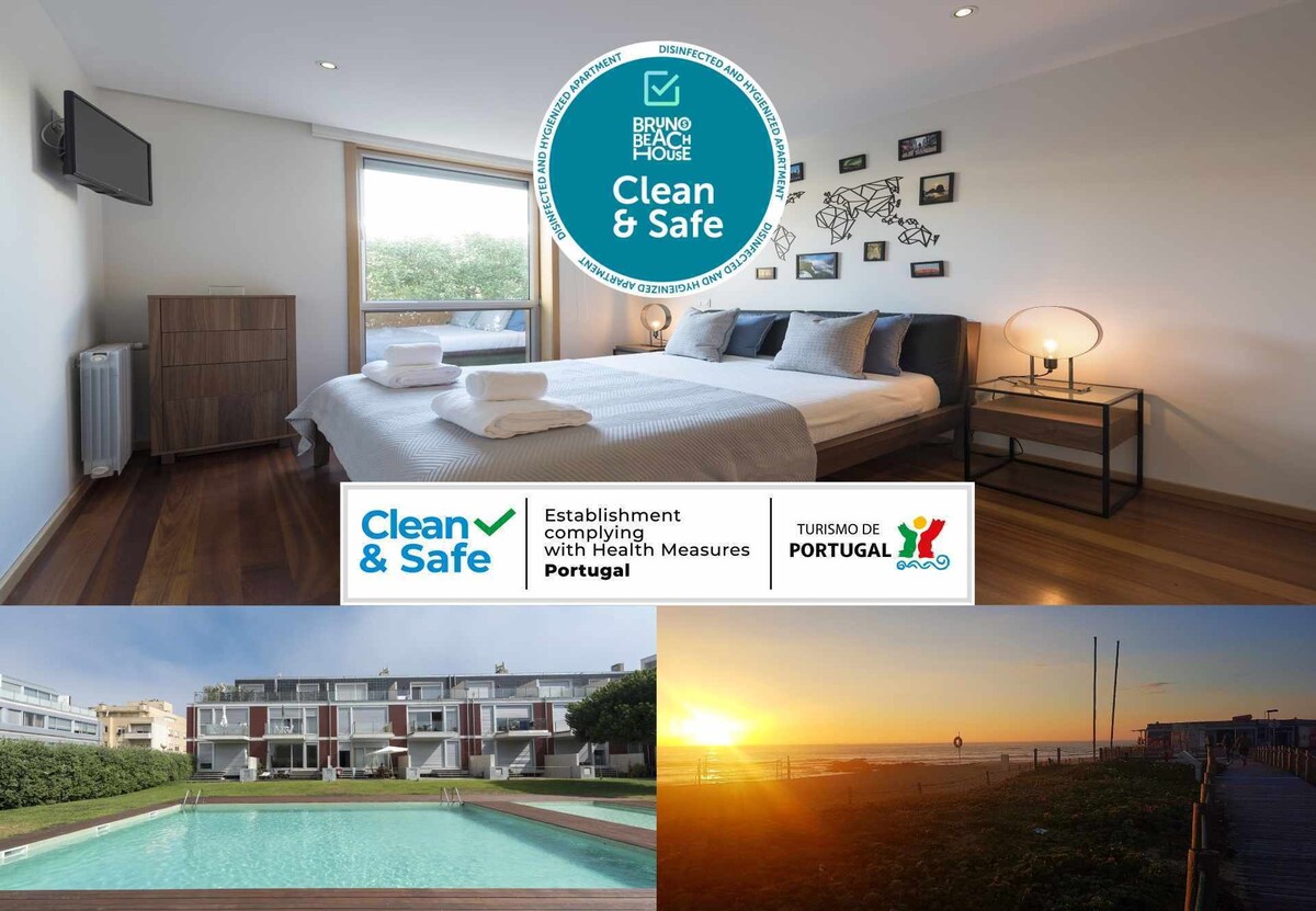 A well-lit bedroom features a spacious queen-size bed, complemented by two pillows and a bedside table with a lamp. A flat-screen TV is mounted on the wall, and large windows offer views of the surroundings. A glimpse of the outdoor pool and ocean can be seen in the lower section of the image.