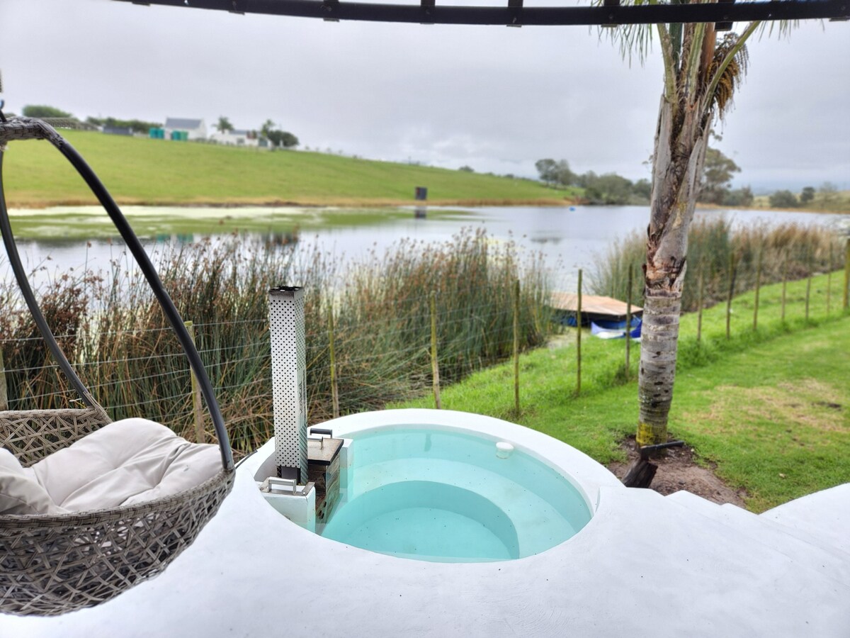 A serene outdoor area features a circular wood-fired jacuzzi surrounded by lush greenery. A comfortable hanging chair provides a cozy spot to relax, while a calm dam reflects the surrounding landscape, enhancing the peaceful atmosphere of this unique retreat.