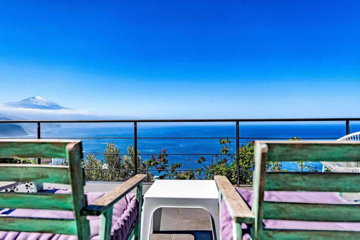 Two lounge chairs are positioned on a private terrace, providing a clear view of the sea and the distant Pico del Teide volcano. A small table is placed between the chairs, complemented by blooming greenery along the balcony's edge under a bright blue sky.