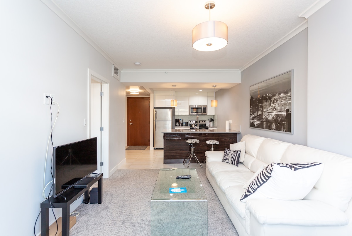 The modern living area features a comfortable white sofa positioned beside a glass coffee table. A flat-screen TV is located on a black stand, and the open kitchen is visible in the background, showcasing stainless steel appliances and a dining bar.