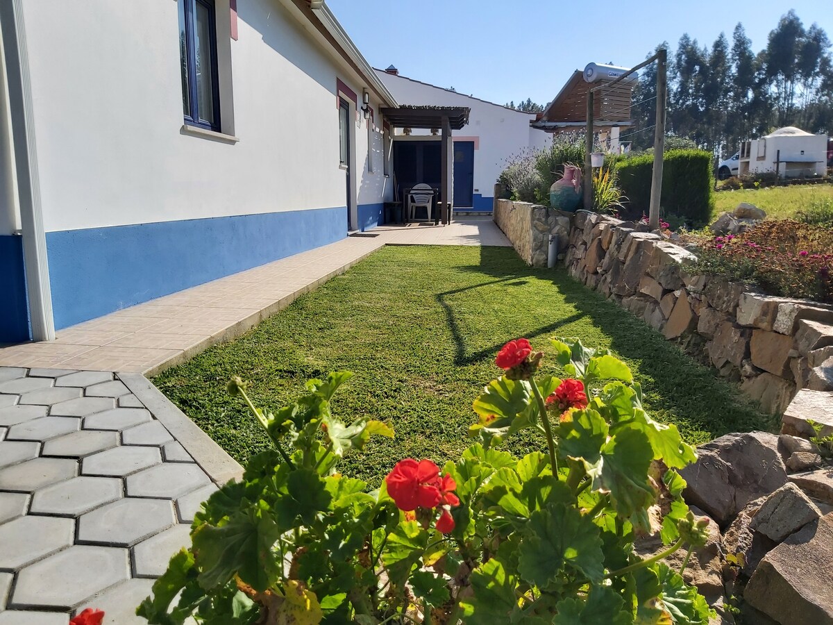 A well-maintained garden is visible in the foreground, featuring blooming red flowers. The tidy lawn is bordered by a stone wall, leading to a patio area with outdoor seating. The house presents a bright exterior with blue accents, offering a welcoming appearance.