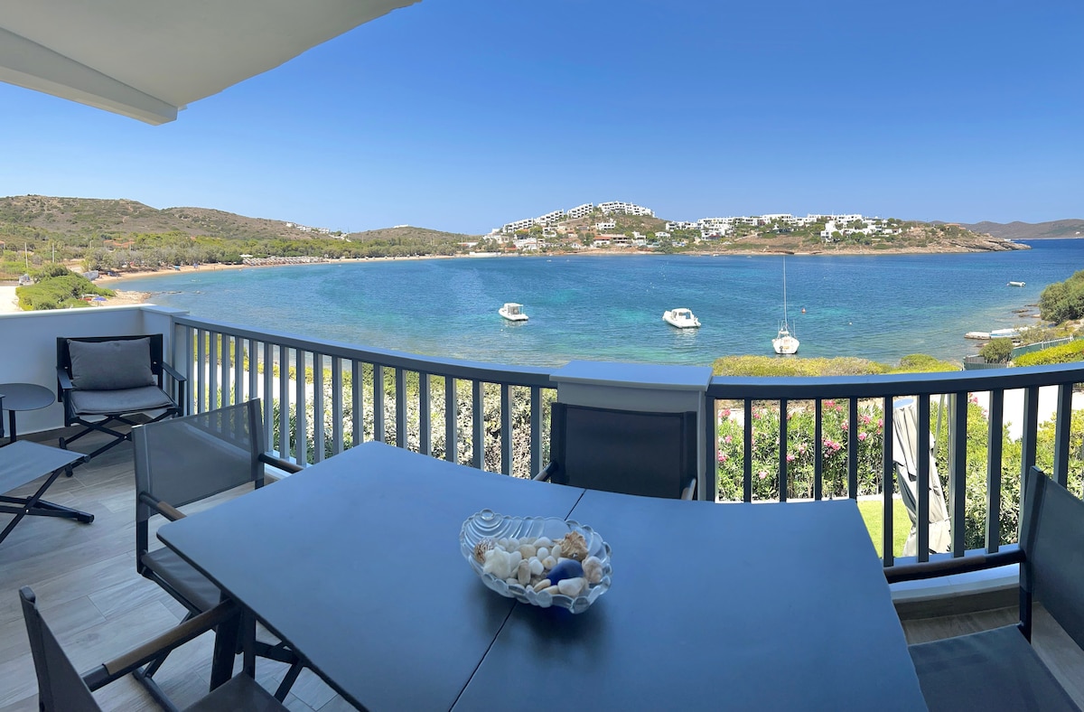 A spacious balcony is presented with a table and chairs, overlooking a serene bay. Boats are anchored in the calm water, and lush greenery surrounds the area. Clear blue skies and a distant shoreline enhance the inviting outdoor space.
