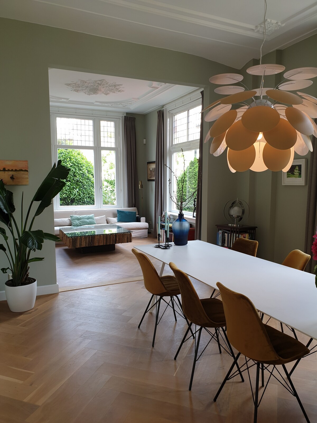 A dining area features a long white table surrounded by modern mustard-colored chairs. A stylish pendant light hangs above. In the background, large windows provide natural light to a cozy living space with a plush sofa and greenery visible outside.