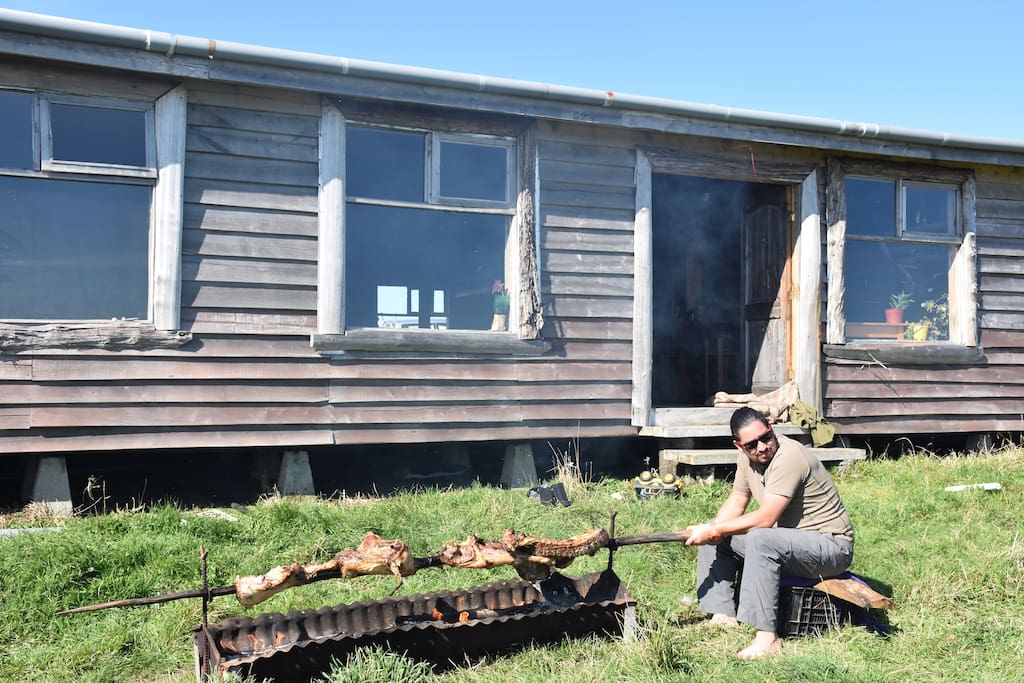 El frontis del hogar con un día de sol, el hermano mayor de la familia, haciendo un asado de cordero al palo, una tradición de la patagonía insular chilota.