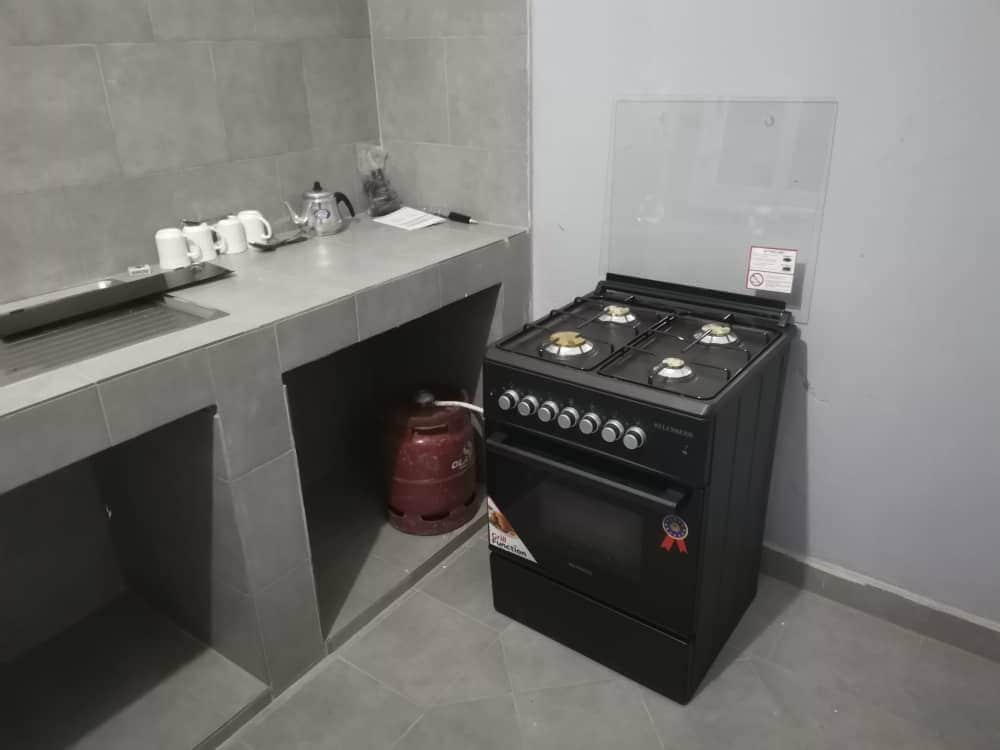 A modern kitchen area features a black gas stove with four burners, positioned next to a gray countertop. A gas cylinder is visible beside the stove, and utensils are neatly arranged nearby, ensuring a functional cooking space.