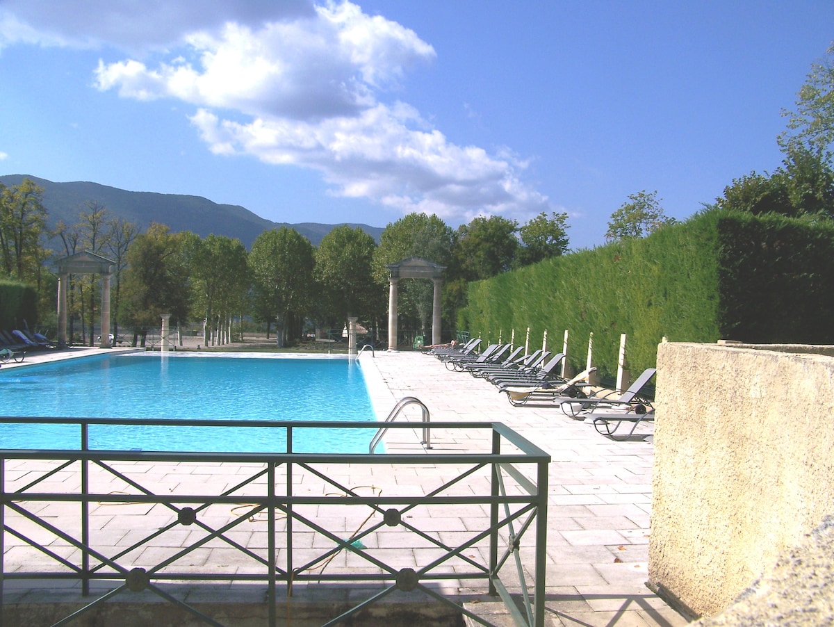 A large swimming pool is featured, bordered by a spacious patio with sun loungers. Lush greenery surrounds the area, providing a sense of privacy. Views of distant mountains are visible in the background, highlighting the serene environment.