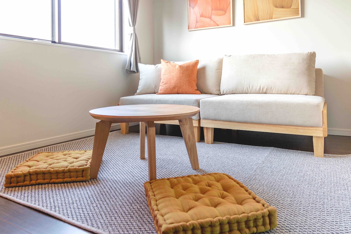 A cozy living area features a light-colored sofa with an accent pillow. A round wooden table is positioned in front, surrounded by textured floor cushions on a woven rug. Natural light streams through a large window with soft curtains.