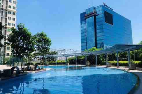 A large swimming pool is surrounded by lush greenery, with tall trees and manicured bushes lining the area. The building's reflective glass facade and modern architecture are visible in the background, emphasizing the urban ambiance.