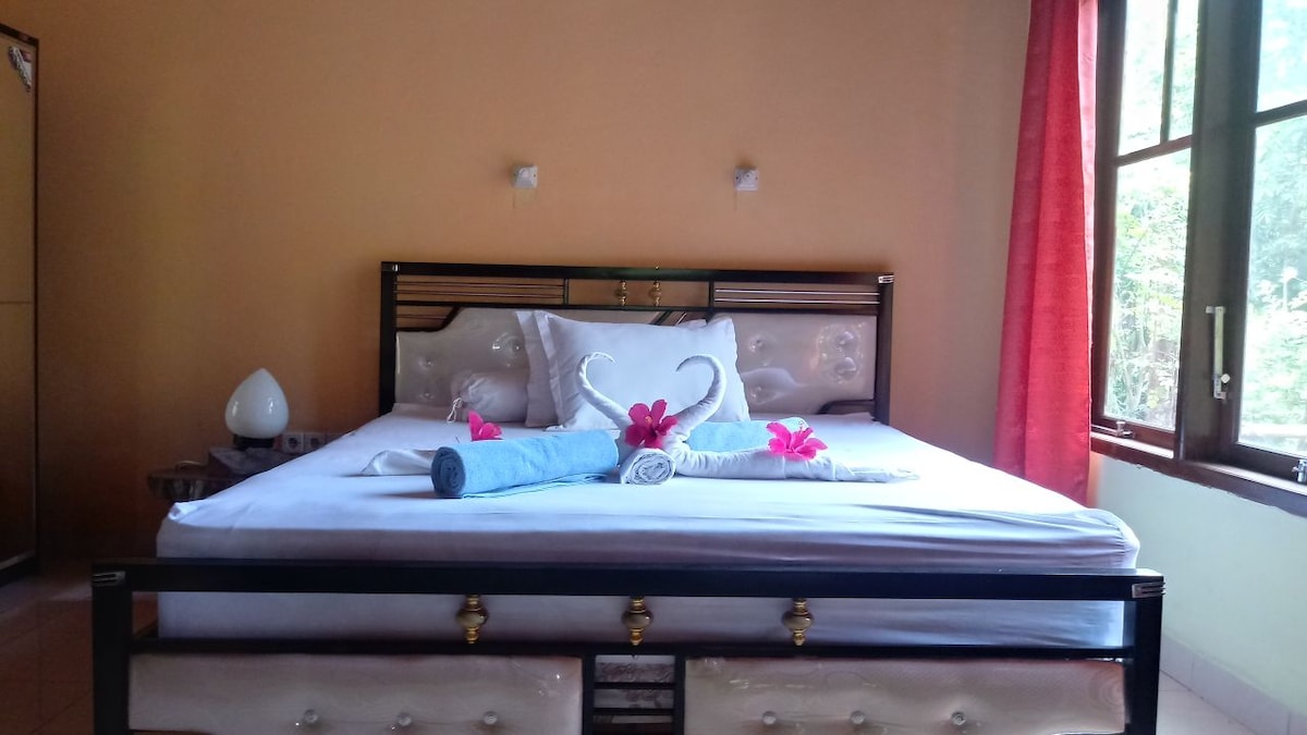 A spacious bed is framed in a decorative headboard, adorned with towels arranged into swan shapes. Delicate flowers add a touch of color. A wooden bedside table holds an elegant lamp. Large windows allow natural light to fill the room, complemented by airy orange walls.