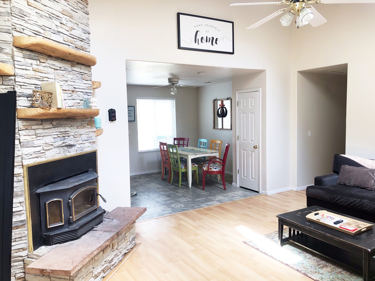A spacious living area is enhanced by a stone fireplace and a cozy sofa. A colorful dining table with mixed chairs is visible in the adjacent room, bathed in natural light from a nearby window. The warm and inviting atmosphere is suited for relaxation.