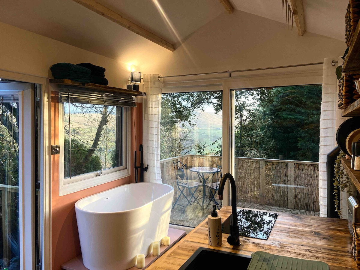 A bright bathroom space features a free-standing bathtub positioned next to expansive windows, offering views of the surrounding hills. Warm lighting enhances the natural wood accents on the countertop and sink, while outdoor seating is visible on the balcony through the glass doors.