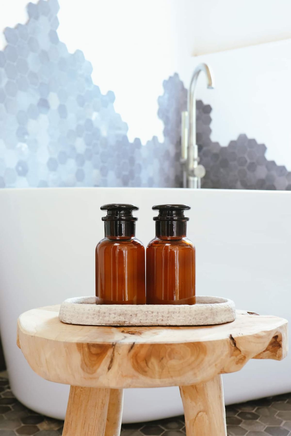 Two amber glass bottles are placed on a natural wooden stool, complementing a modern freestanding bathtub in the background. The wall features a unique hexagonal tile pattern in varying shades of gray, enhancing the space's contemporary design.
