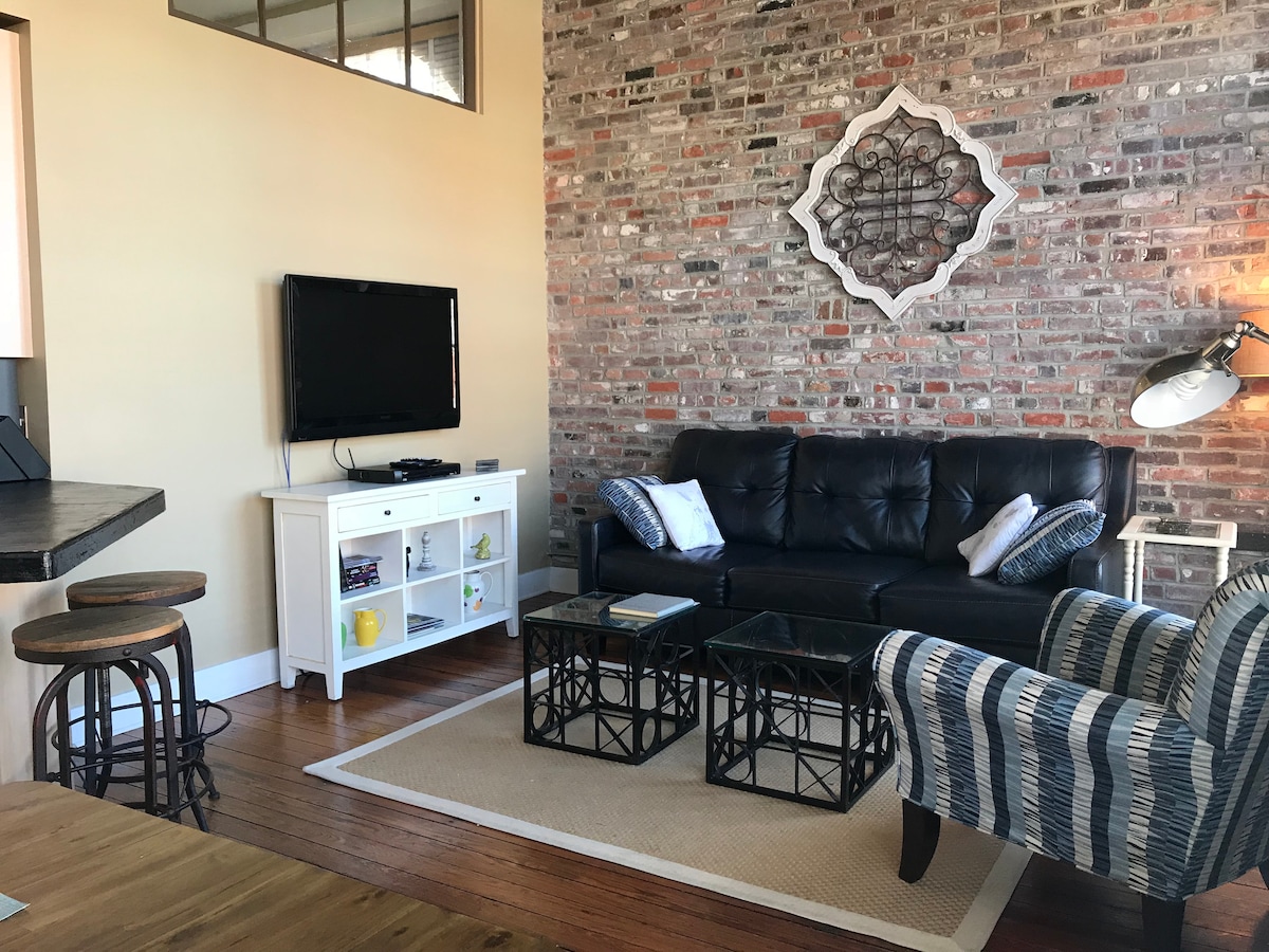 A living area features exposed brick walls, creating an industrial yet cozy ambiance. A leather sofa is paired with a striped accent chair, while a coffee table with glass tops sits on a woven area rug. A flat screen TV is mounted on the wall, providing entertainment options.