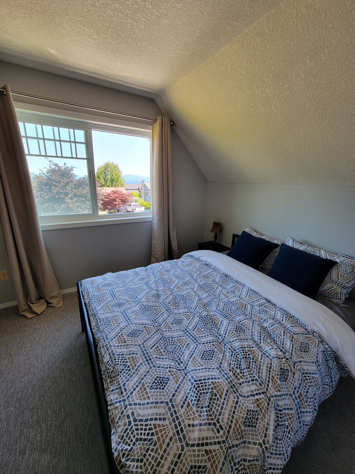 A well-lit bedroom features a queen bed with a patterned duvet and two blue accent pillows. Natural light filters through a large window, adorned with beige curtains, which offers a view of the outdoors. The serene color scheme and soft carpeting enhance the inviting space.