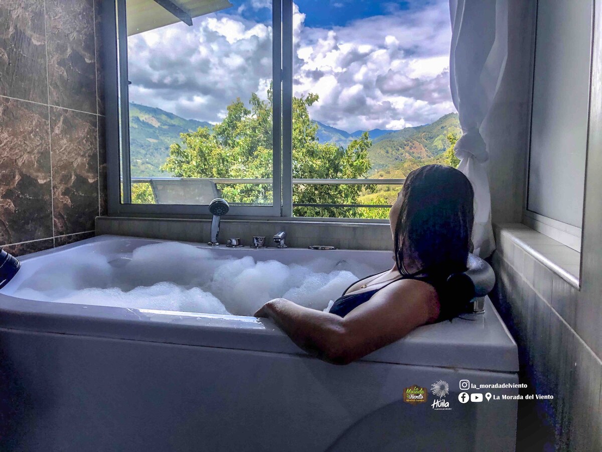A modern bathroom features a private jacuzzi tub positioned by a large window. Views of the surrounding mountains and cloudy skies are visible, while bubbles fill the tub. Natural light illuminates the serene space, enhancing the sense of tranquility.