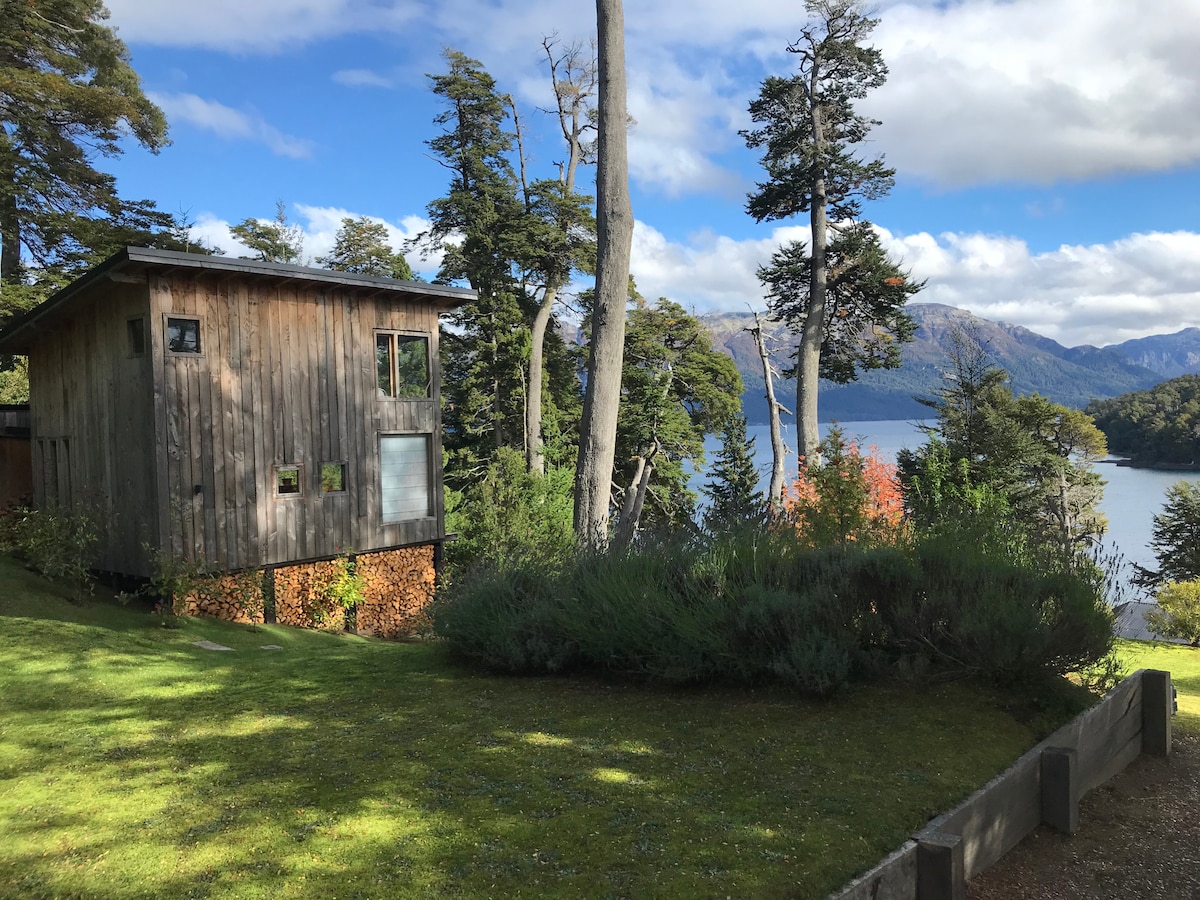 A wooden cabin stands surrounded by lush greenery, with a view of the tranquil Nahuel Huapi Lake and mountains in the background. Tall trees provide shade, and logs are stacked neatly underneath the cabin. The sky displays a mix of clouds and blue patches.