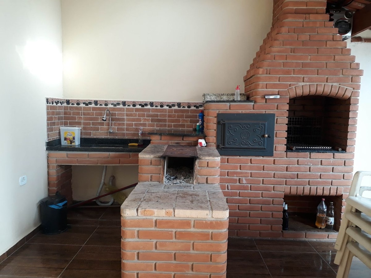 An outdoor kitchen space is showcased, featuring a traditional brick barbecue grill and an integrated stove. A countertop for food preparation is included beside a sink, with a firewood storage section visible. The area is well-lit, enhancing functionality and promoting outdoor cooking.