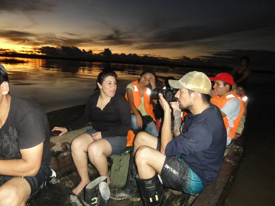 A group of individuals is seated on a boat during twilight, surrounded by calm waters reflecting a colorful sunset. Some wear life jackets, and one person is seen capturing the scene with a camera. The atmosphere suggests an engaging adventure in nature.