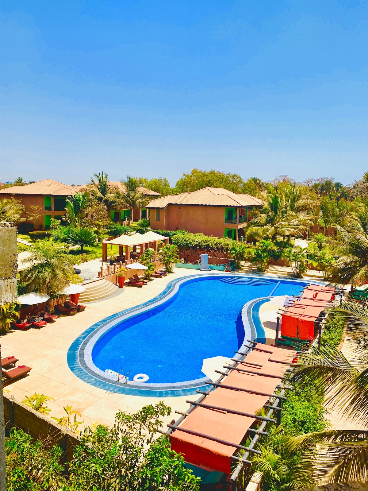 An inviting swimming pool with a curvy design is surrounded by lush palm trees and comfortable lounge areas. Several shaded cabanas provide seating options, while umbrellas stand near the pool's edge. The clear blue water reflects the bright sky, creating a peaceful atmosphere.