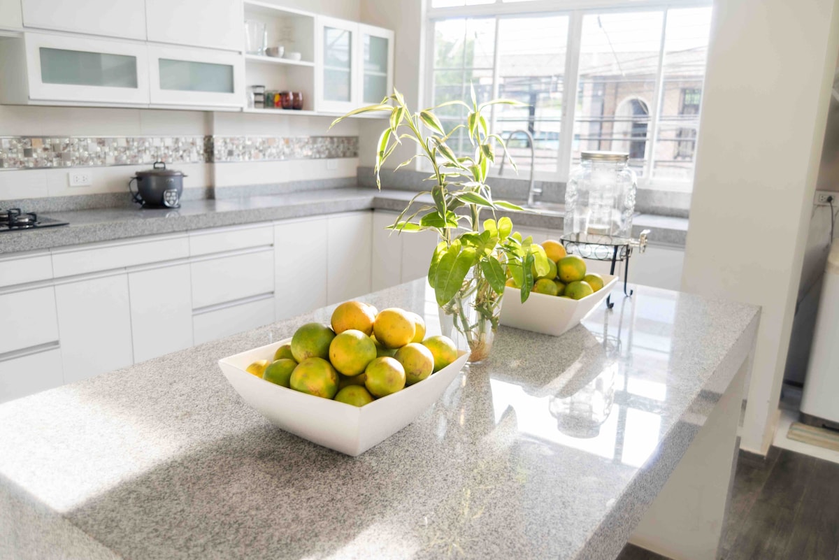 A modern kitchen features sleek white cabinetry and a spacious granite countertop. Natural light pours in from a large window, illuminating a centerpiece comprising bowls filled with fresh lemons and limes. A small plant adds a touch of greenery to the space.