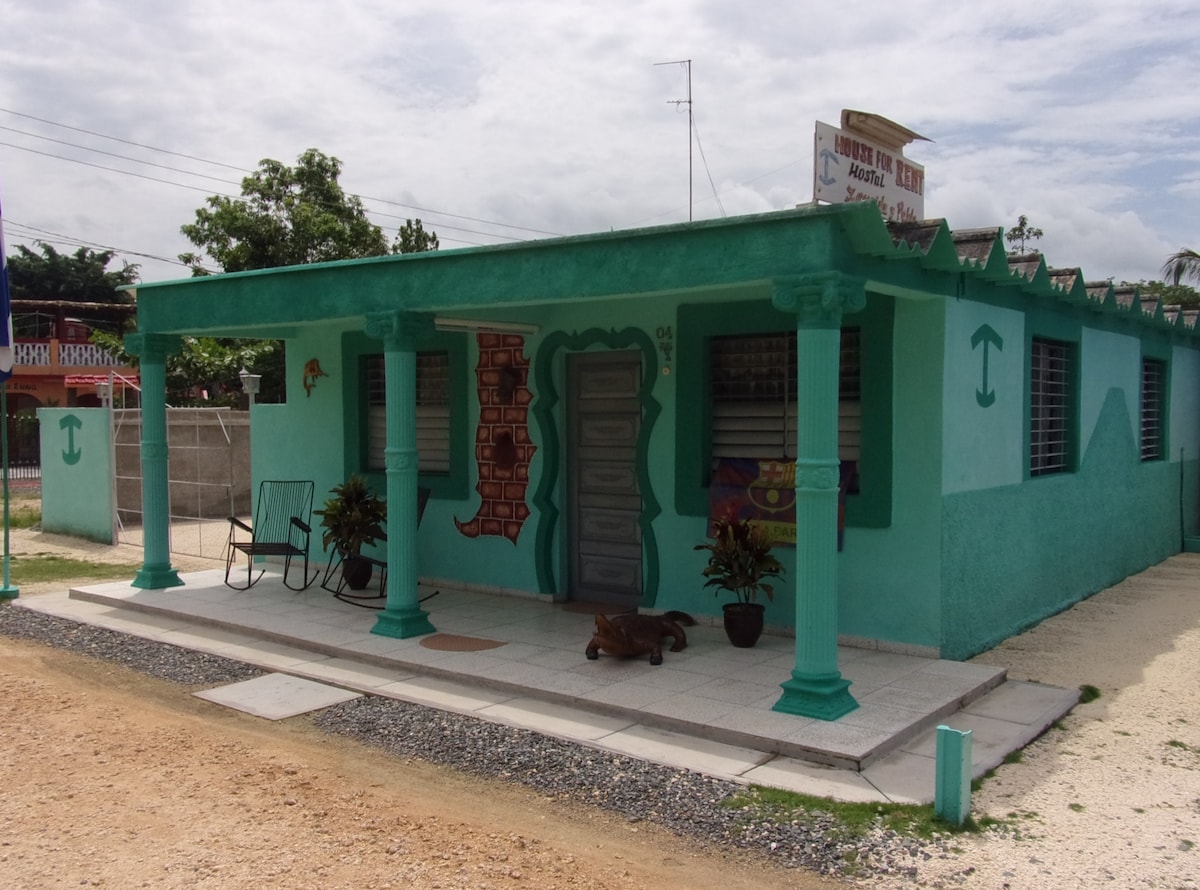 A colorful exterior of a single-story building is displayed, featuring teal walls and decorative accents. A covered porch is supported by two pillars, with a chair positioned nearby. Small plants and artwork add charm to the entrance, complemented by a gravel pathway.