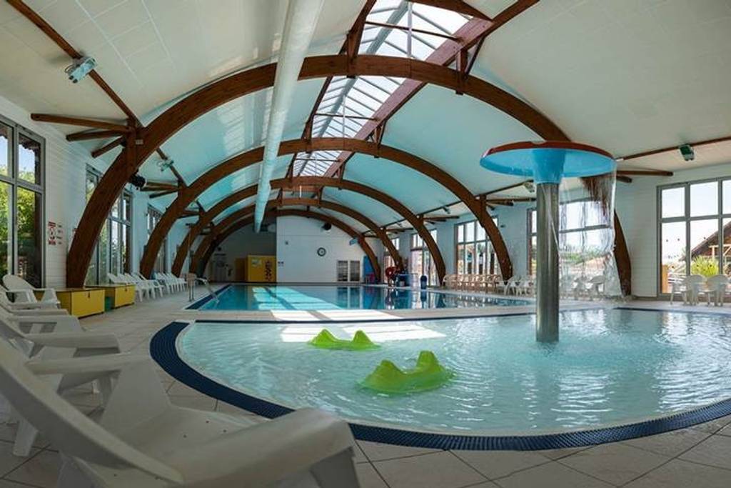 An indoor pool area is highlighted by a large swimming pool, complemented by a children’s splash zone featuring playful water structures. Sunlight enters through skylights and large windows, illuminating wooden beams overhead. Several white lounge chairs are positioned around the pool's perimeter.