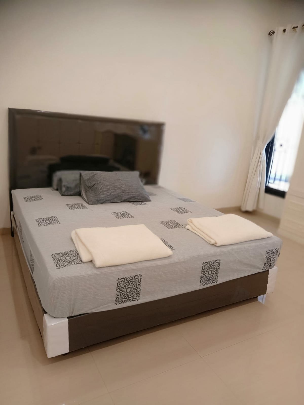 A spacious bed is dressed in a light gray coverlet featuring subtle patterns, with two neatly folded towels placed on top. A single pillow rests against the headboard, and natural light enters through a nearby window adorned with soft curtains.