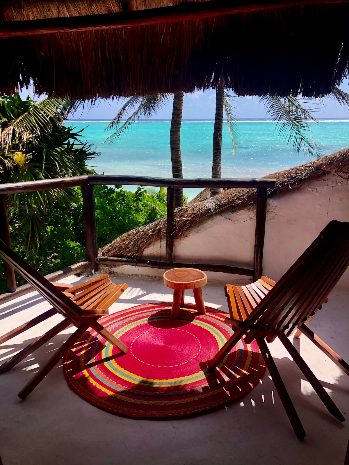 A private balcony is shown with two wooden chairs facing the ocean, accompanied by a small circular table. A colorful round rug enhances the space, while palm trees and a thatched roof create a tropical ambiance against the clear blue waters of the bay.