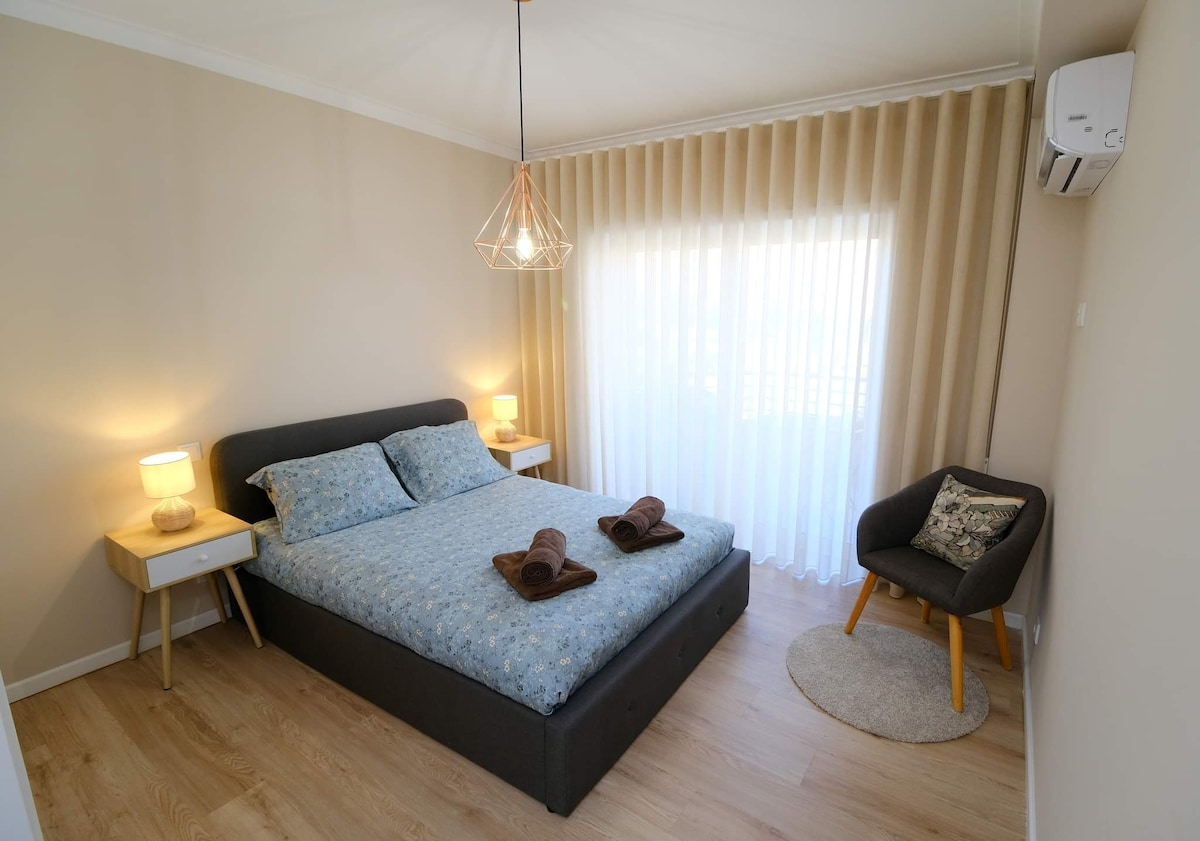 A serene sleeping space is arranged with a spacious bed featuring a floral blue duvet. Two bedside lamps provide soft lighting, and a comfortable armchair is positioned nearby. Sheer curtains filter light from the window, enhancing the room's calming atmosphere.