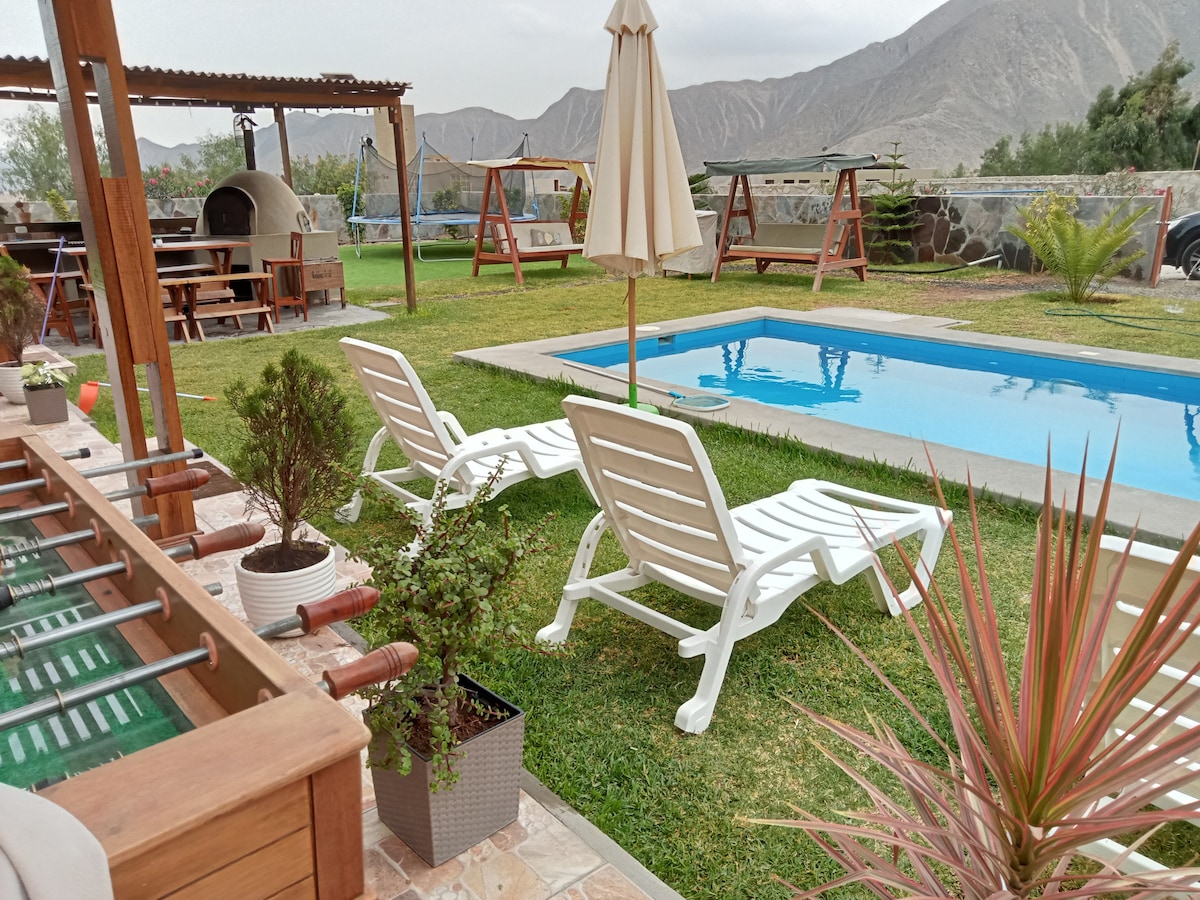 A private pool is surrounded by well-maintained grass, with two lounge chairs facing the water. Nearby, a rustic dining area is visible, featuring a clay oven and a shade umbrella. Mountains form a scenic backdrop, enhancing the tranquil outdoor space.