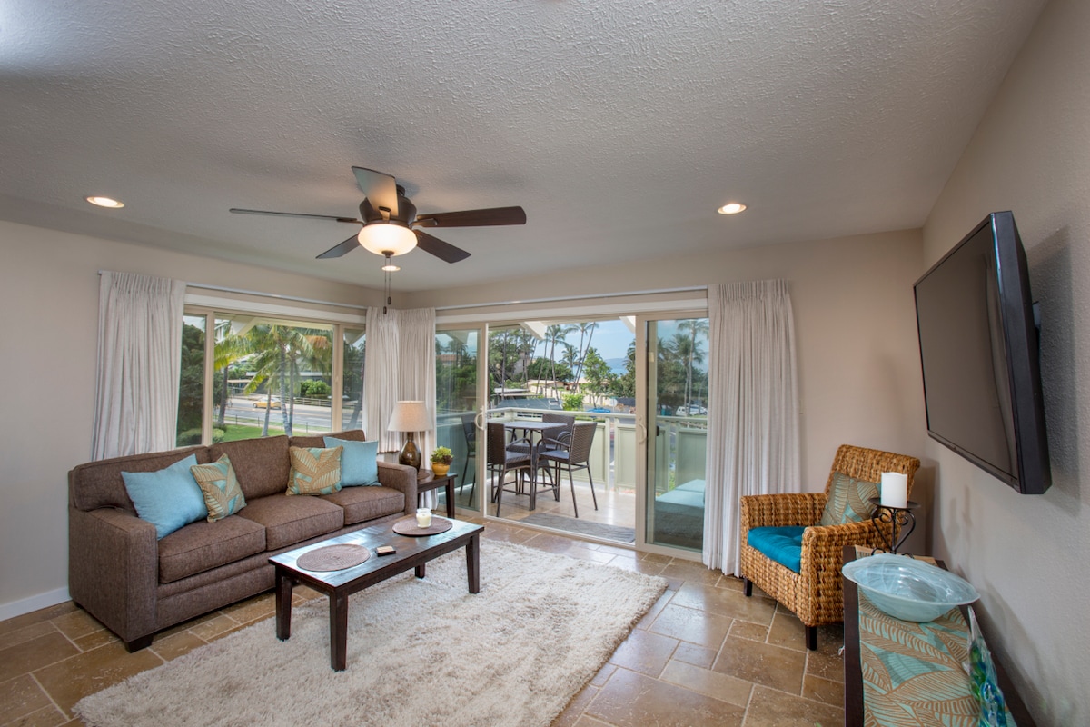 The living area features a comfortable sofa adorned with decorative cushions. A coffee table sits centrally on a soft rug, and large glass doors provide views of the outdoor space. Natural light fills the room, complemented by a ceiling fan and mounted television.