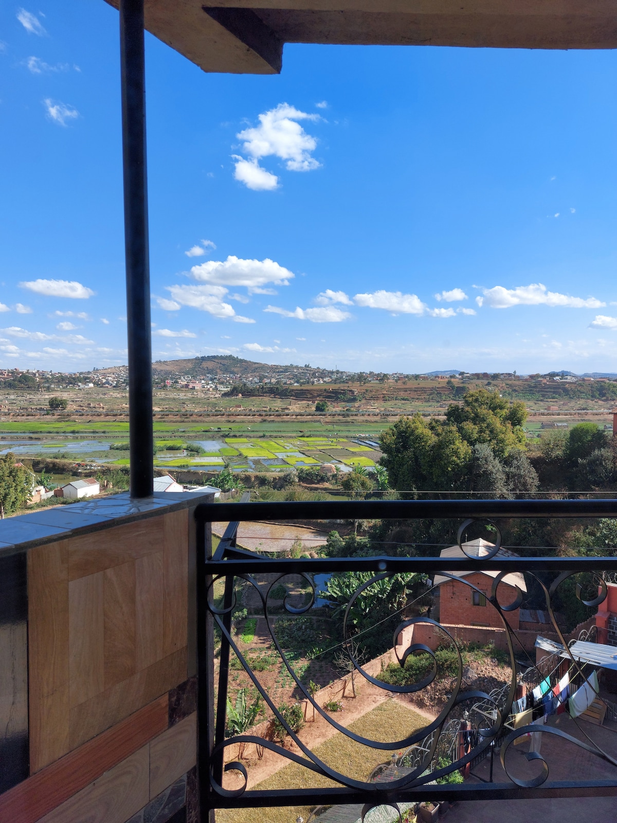 A balcony view showcases a sweeping landscape, including lush fields and distant hills under a bright blue sky with scattered clouds. Decorative railing adds a touch of elegance, while trees and structures are visible in the foreground, blending with the serene surroundings.