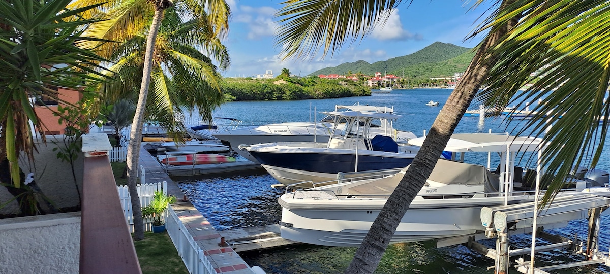A scenic view of the lagoon showcases several boats docked at a private area, framed by lush palm trees. The water reflects the clear blue sky with wispy clouds and hillside greenery in the background, providing a peaceful and serene environment.