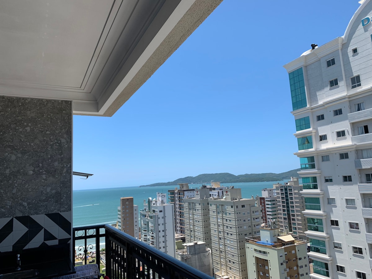 A balcony view is presented, highlighting a blend of city buildings and the ocean beyond. The clear blue sky and calm sea create a serene backdrop, while neighboring structures reflect urban life. The outdoor space features a portion of the balcony's railing, framing the expansive view.