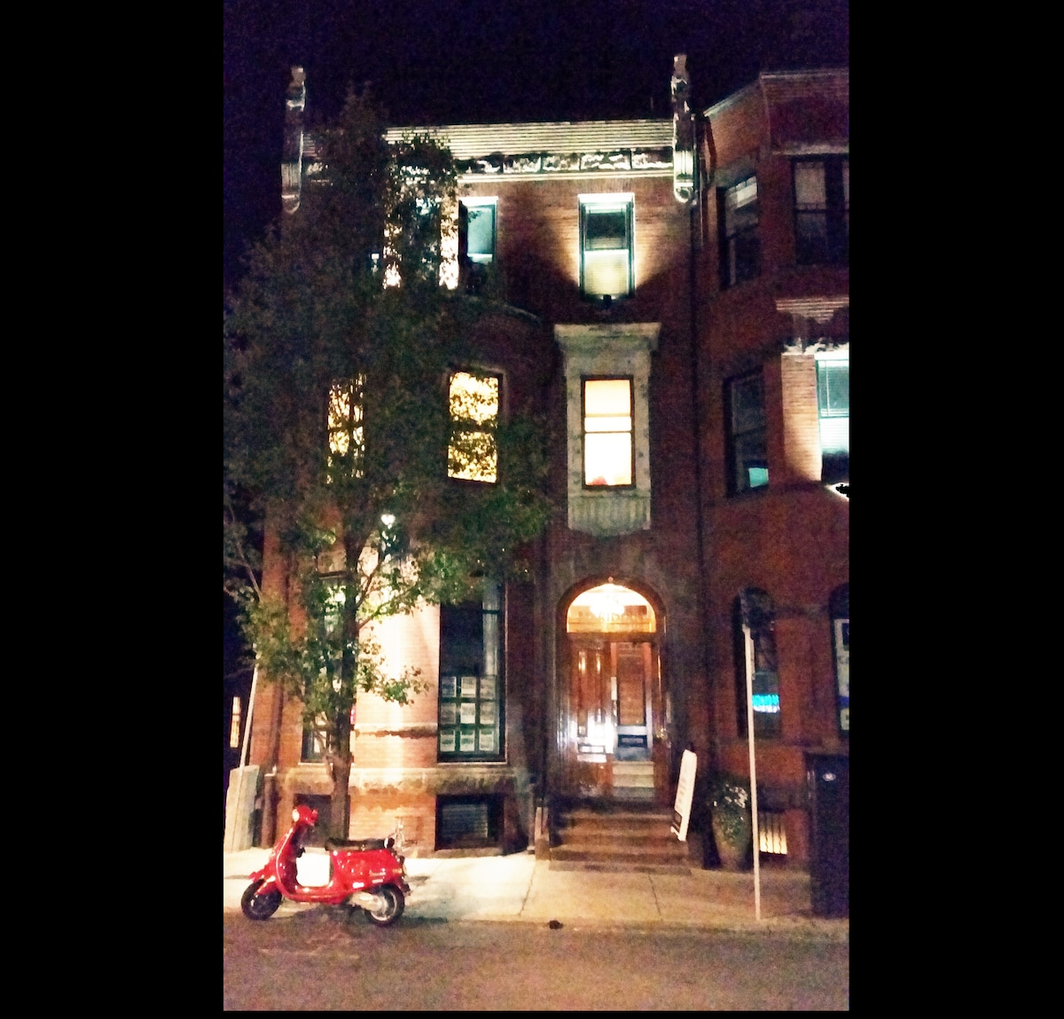 The historic brownstone building is illuminated at night, showcasing its architectural details. A warmly lit entrance is flanked by large windows, while a red scooter is parked at the curb. The surrounding street is quiet, with minimal noise visible.