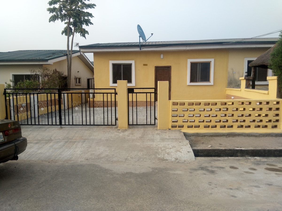 A single-story yellow house is framed by a low brick wall and a metal gate, providing a welcoming entrance. A satellite dish is visible on the roof. The surrounding area features greenery, and a nearby building can be glimpsed in the background.