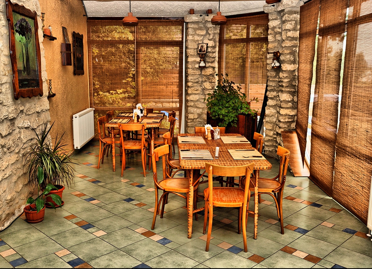 A dining area is showcased with natural stone walls and wood accents. Multiple wooden tables and chairs are arranged, each featuring a simple table setting. Large windows allow for ample natural light, complemented by woven blinds. Potted plants add a touch of greenery to the space.
